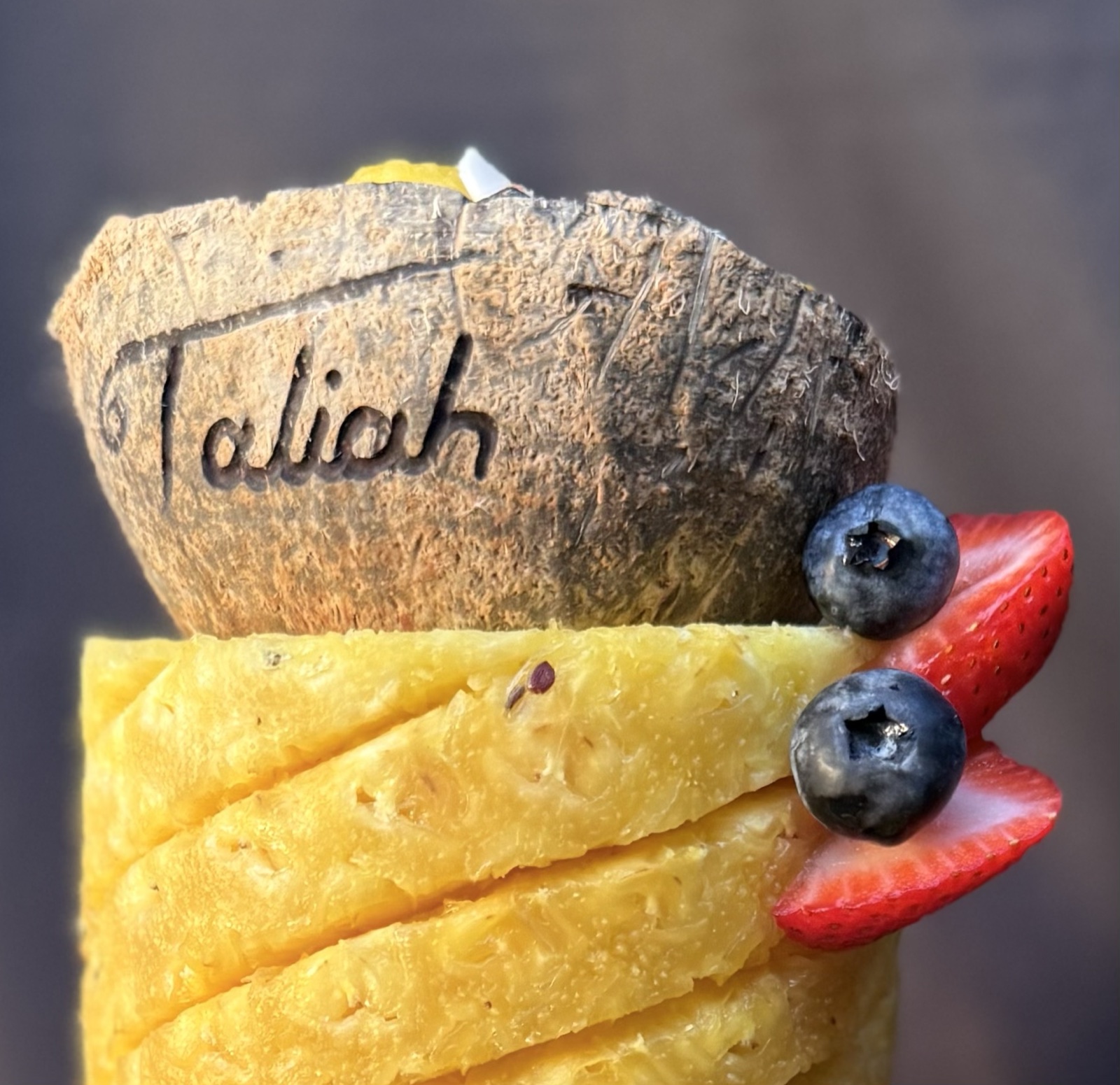 Coconut shell hand-engraved with the name Taliah on top of a pineapple cake with strawberries and blueberries