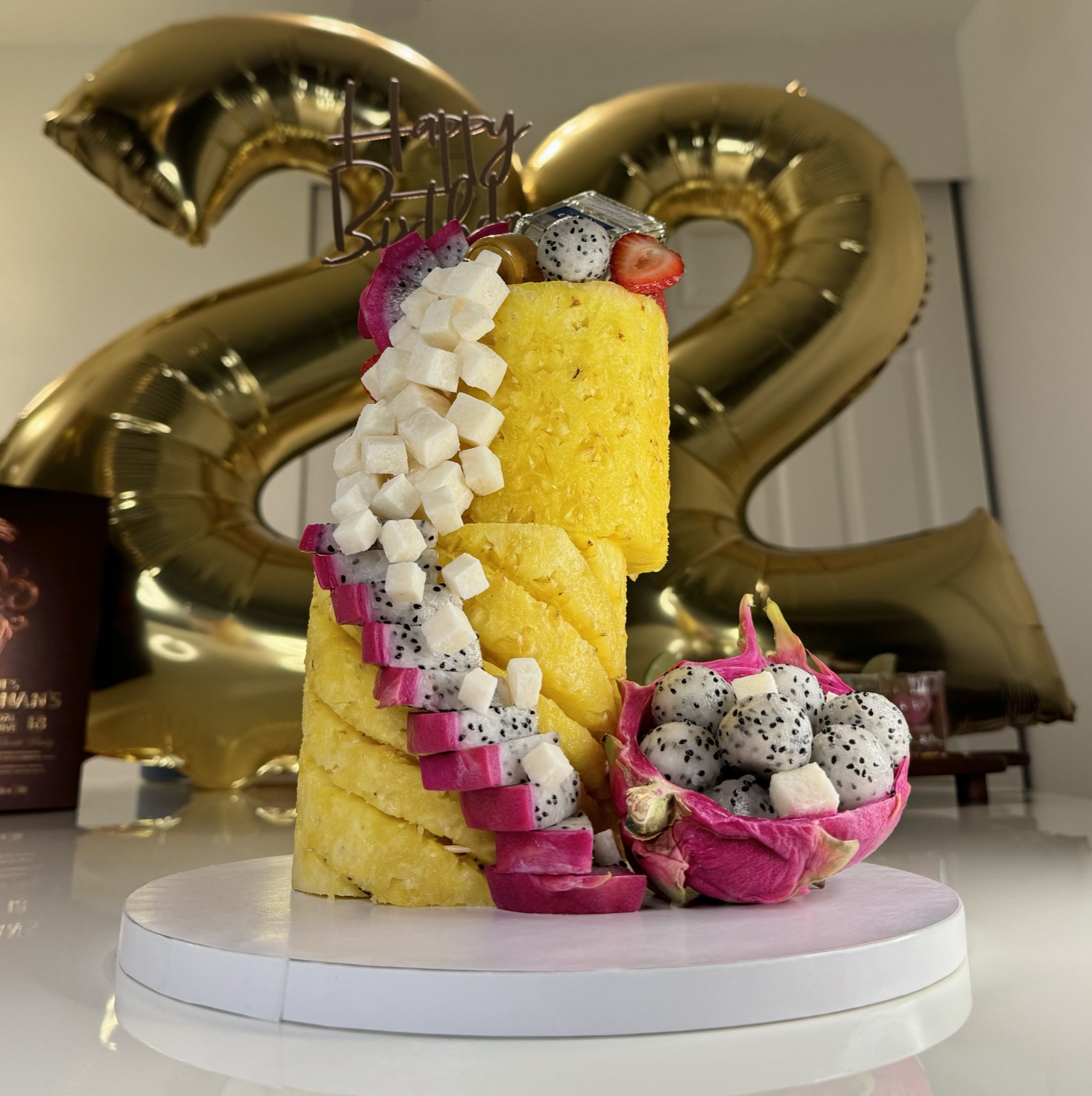 Two-tier pineapple cake with dragon fruit and j&iacute;cama with a Happy Birthday topper and gold balloons